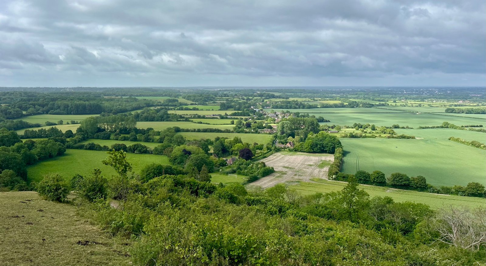 Panoramic view of the Kent countryside with rolling green fields, woodlands, and scattered farmhouses under a cloudy sky.