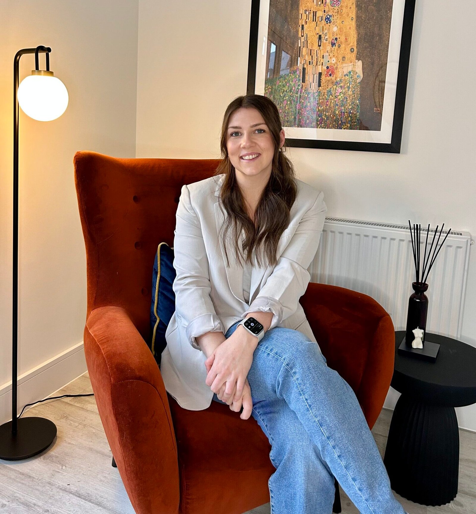 Smiling woman seated in a burnt orange armchair with folded legs, wearing a blazer and jeans, in a cosy modern interior.