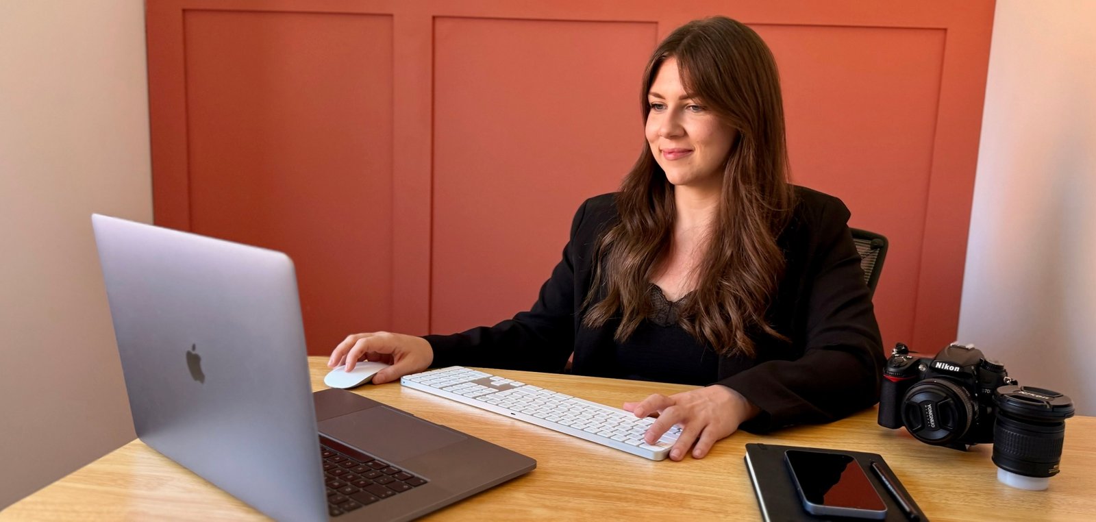 Gabbi, founder of Ace Marketing, working at her desk with a MacBook, external keyboard, and camera equipment in a warm-toned office.