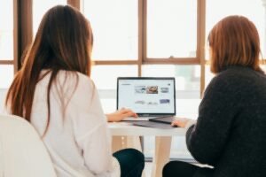 Two women sitting at a table reviewing a website on a laptop in a bright office space with large windows.