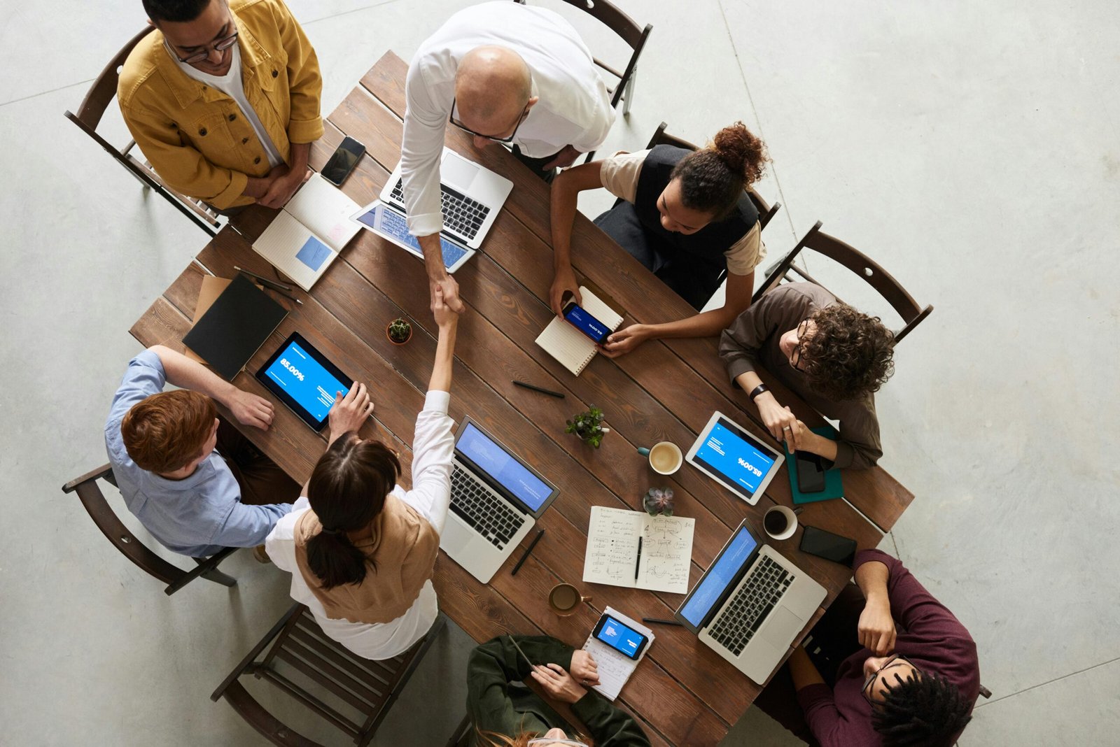 Top-down view of a diverse team gathered around a large wooden table with laptops, tablets, notebooks and coffee, as two people shake hands in the centre.
